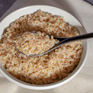 A bowl of cooked brown rice with a spoon, surrounded by uncooked grains.